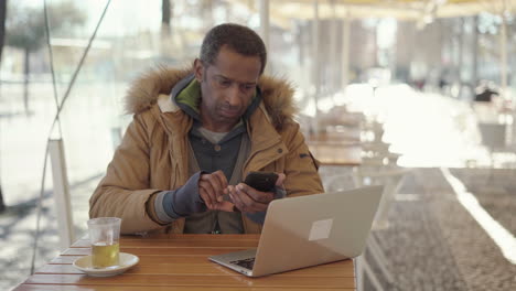 Hombre-Afroamericano-Usando-Laptop-Y-Teléfono-Celular-En-La-Cafetería